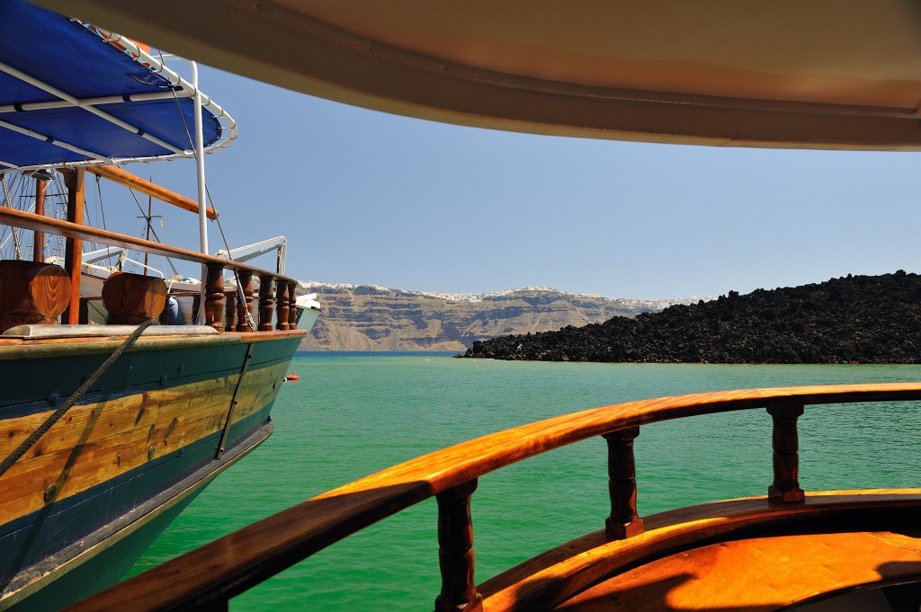 Kykladen-062009-159.jpg - Blick unter Deck aus dem Heck der "Albatross" in Richtung des Hafens von "Nea Kameni". Das Wasser ist wegen der vulkanischen Aktivität so unnatürlich grün. Der schwarze Schutthaufen ist das Ende eines Lavastroms der hier beim letzten größeren Ausbruch erstarrt ist. Neben uns liegt noch ein weiteres Ausflugsboot gleichen Typs.